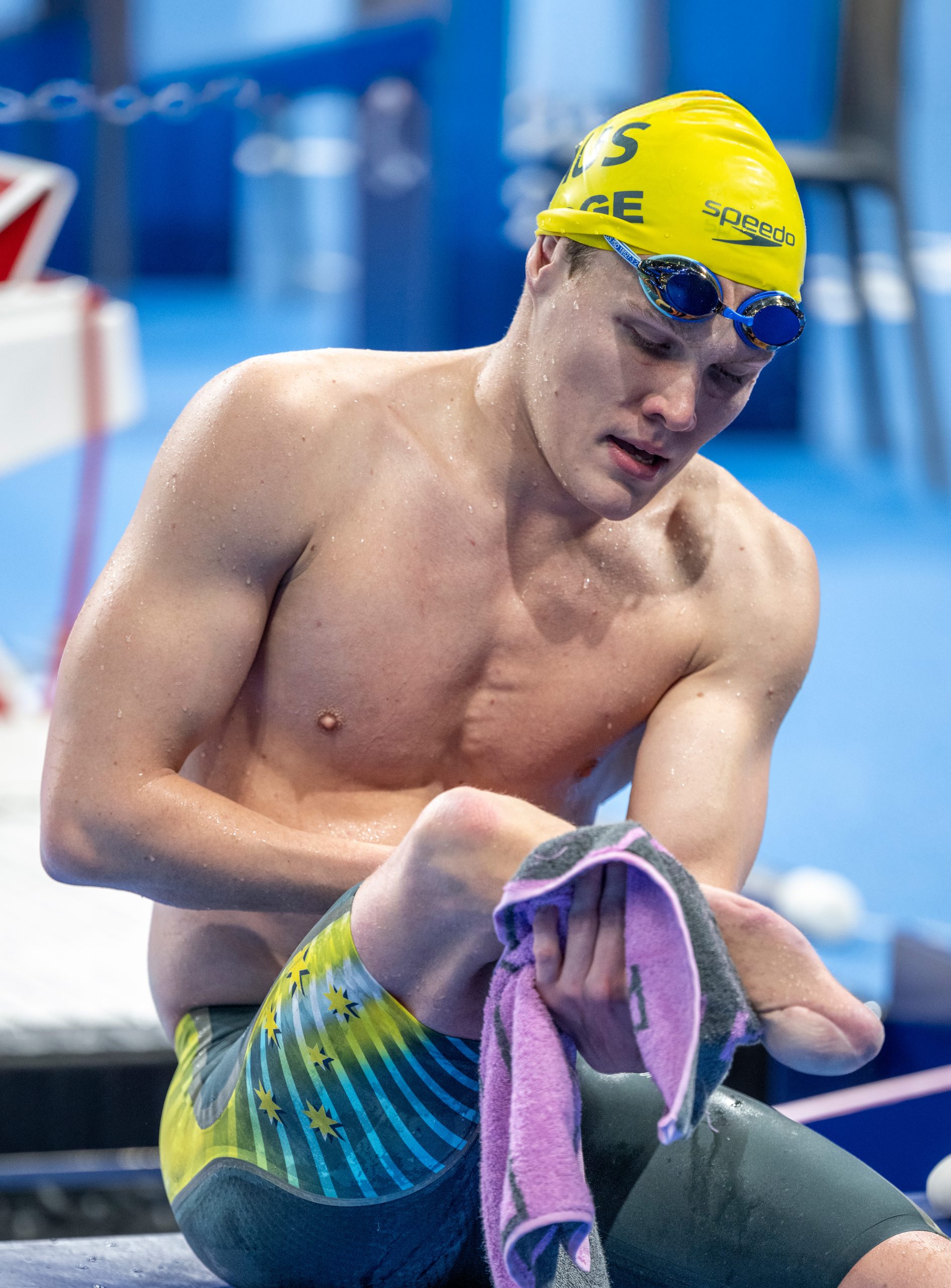 A male para swimmer wearing a bright yellow swim cap wipes his amputee leg with a blue and purple towel. He sits poolside in swim trunks, preparing for competition, with a focused expression and aquatic equipment blurred in the background.