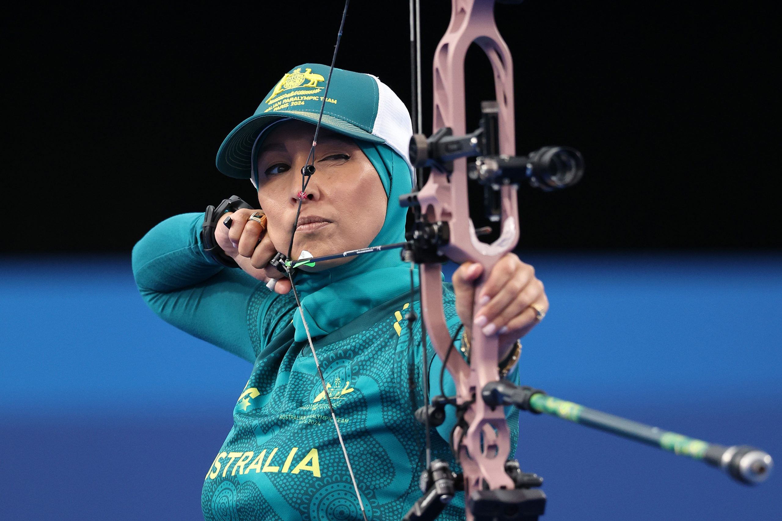 A female para archer dressed in a teal Australia uniform and cap draws back her bow with precision. Her eyes are focused downrange as she prepares to release the arrow. The background is dark, emphasizing her concentration and the intensity of the moment.